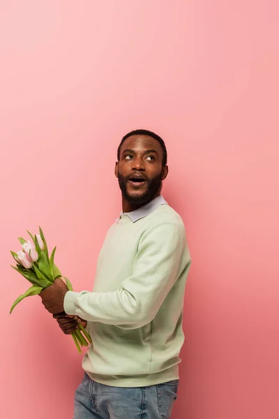 Erstaunt afrikanisch-amerikanischer Mann schaut weg, während er Tulpen auf rosa Hintergrund hält — Stockfoto