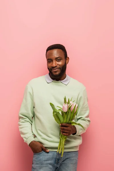 Africano americano hombre con fresco tulipanes sonriendo a cámara en rosa fondo - foto de stock