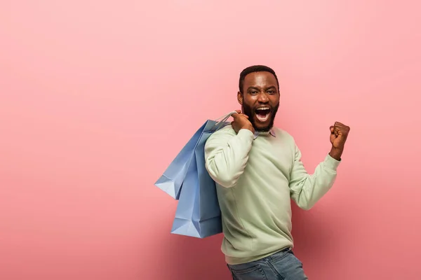 Схвильований афроамериканський чоловік з сумками, що показують радісний жест на рожевому фоні — стокове фото