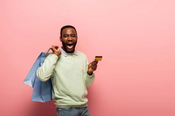 Espantado afro-americano homem segurando sacos de compras e cartão de crédito em fundo rosa — Fotografia de Stock