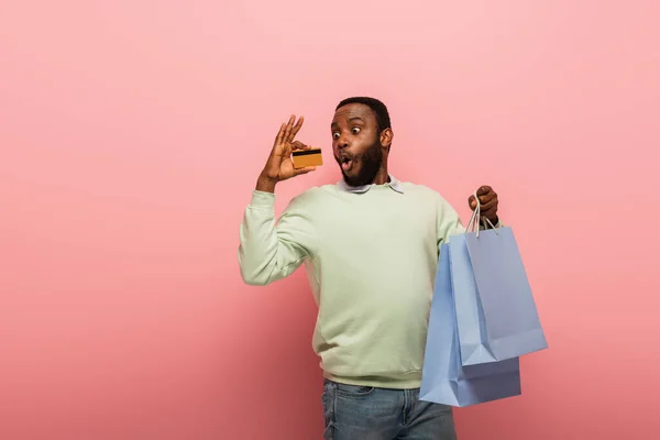 Surpreso homem afro-americano olhando para o cartão de crédito, enquanto segurando compras em fundo rosa — Fotografia de Stock