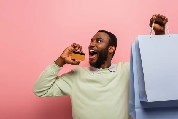 Hombre afroamericano emocionado mirando hacia otro lado mientras sostiene la tarjeta de crédito y las compras en el fondo rosa - foto de stock