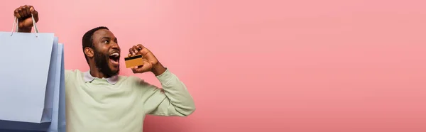 Astonished african american man with credit card and purchases looking away isolated on pink, banner — Stock Photo