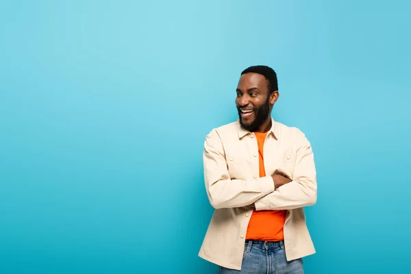 Hombre afroamericano riendo mirando hacia otro lado mientras está de pie con los brazos cruzados aislados en azul - foto de stock