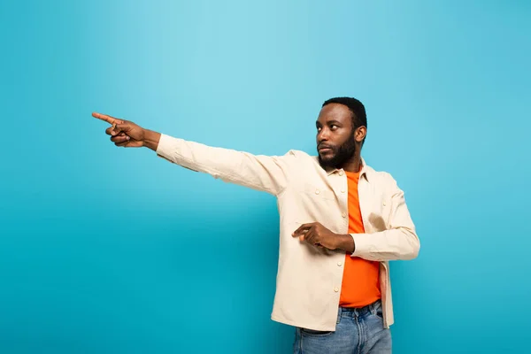 Junger afrikanisch-amerikanischer Mann zeigt mit dem Finger zur Seite auf blauem Hintergrund — Stockfoto