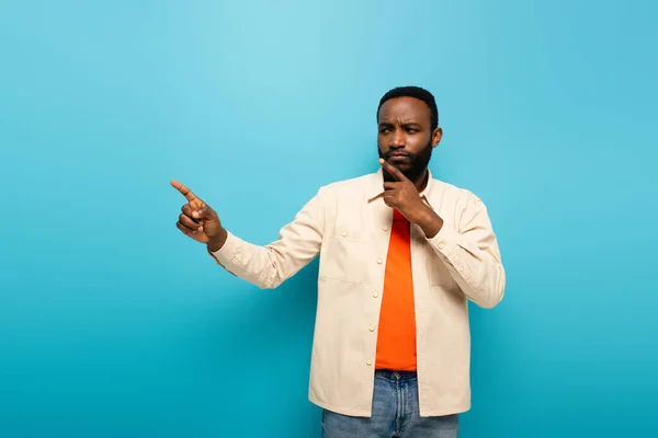 Reflexivo afroamericano hombre tocando la cara mientras señala a un lado con el dedo sobre fondo azul - foto de stock