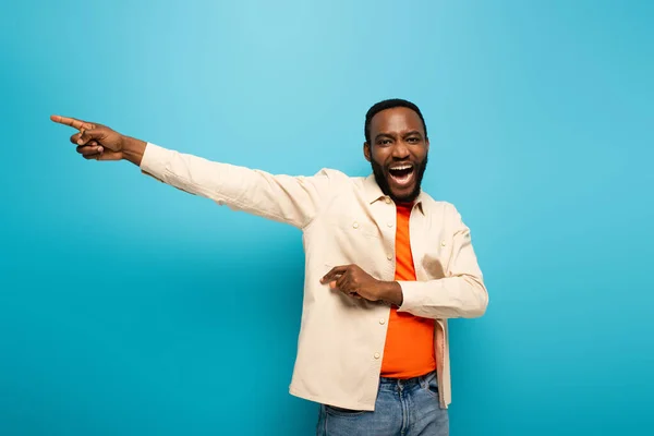 Laughing african american man looking at camera and pointing aside with finger on blue background — Stock Photo