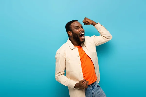 Amazed african american man showing rejoice gesture while looking away on blue background — Stock Photo