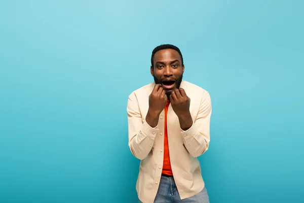 Sorprendido hombre afroamericano agarrado de la mano cerca de la cara mientras mira a la cámara en el fondo azul - foto de stock