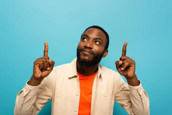 Hombre afroamericano sonriendo mientras señala hacia arriba con los dedos aislados en azul - foto de stock
