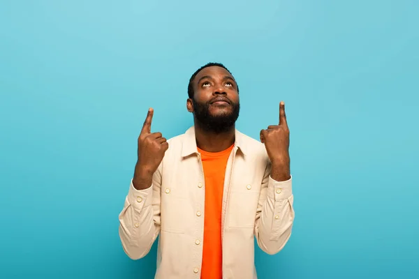 Jovem afro-americano homem olhando para cima e apontando com os dedos isolados em azul — Fotografia de Stock