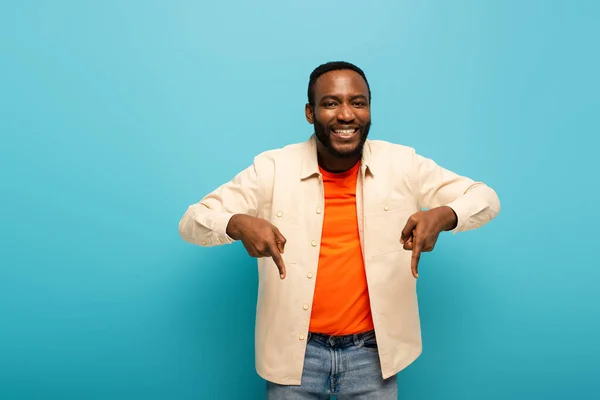 Gioioso afro americano uomo puntando verso il basso con le dita su sfondo blu — Stock Photo