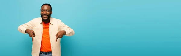 Cheerful african american man pointing down with fingers isolated on blue, banner — Stock Photo