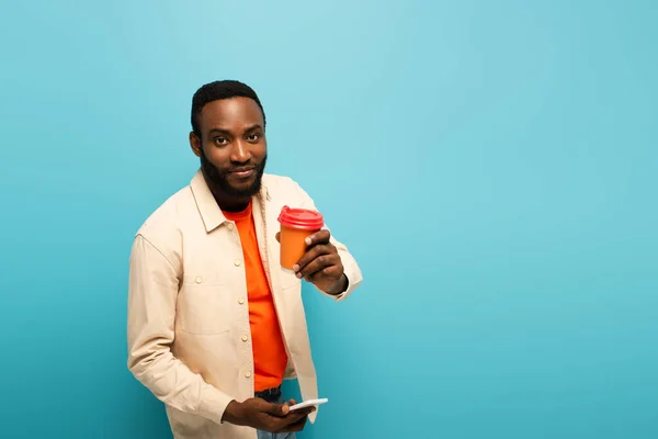 Lächelnder afrikanisch-amerikanischer Mann mit Kreditkarte, während er Handy isoliert auf blauem Grund benutzt — Stockfoto