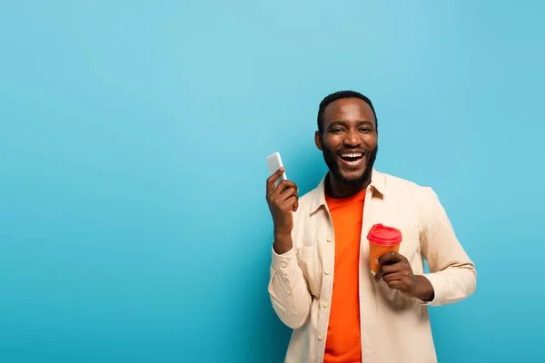 Homem americano africano feliz que prende o cartão de crédito ao usar o telefone móvel isolado no azul — Fotografia de Stock