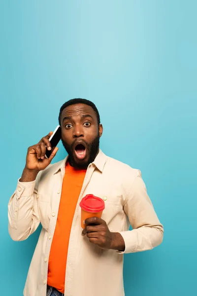 Choqué homme afro-américain avec téléphone portable et café pour aller regarder la caméra isolée sur bleu — Photo de stock