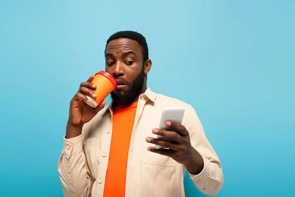 Здивований афроамериканський чоловік п'є каву, дивлячись на смартфон ізольовано на синьому — стокове фото