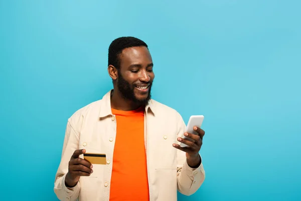 Улыбающийся африканский американец, держащий кредитку, используя мобильный телефон, изолированный на голубом — стоковое фото