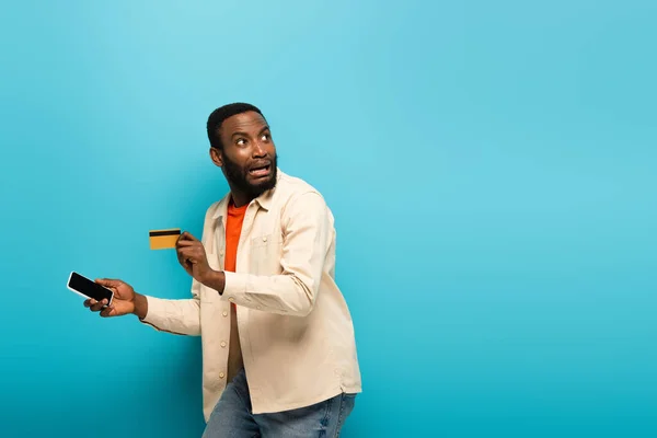 Scared african american man with mobile phone and credit card looking away on blue — Stock Photo