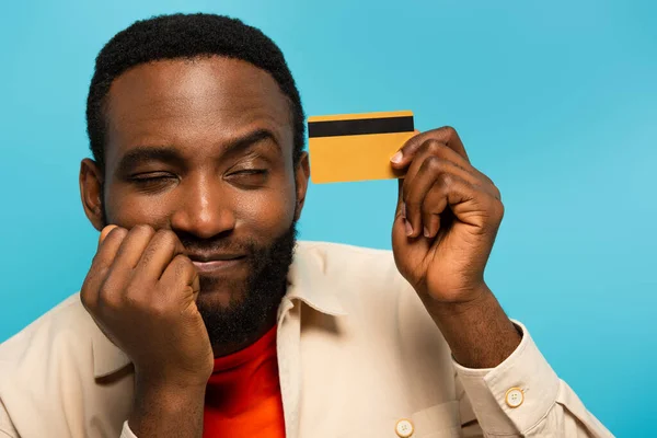 Homme afro-américain positif rêvant tout en tenant la carte de crédit isolée sur bleu — Photo de stock