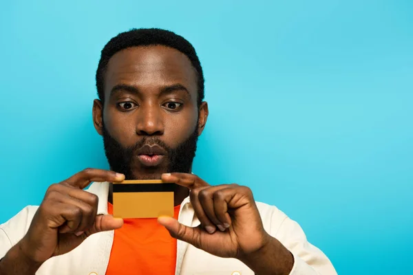 Удивленный африканский американец показывает кредитную карту изолированы на синий — стоковое фото