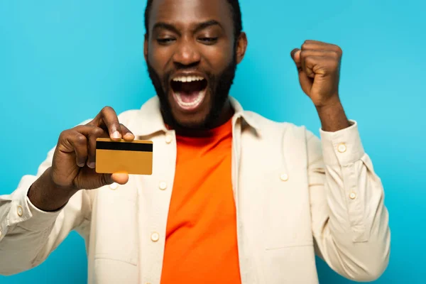 Excited african american man shouting and showing credit card isolated on blue, blurred background — Stock Photo