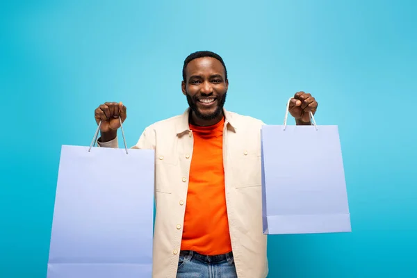 Heureux homme afro-américain souriant à la caméra tout en tenant des achats isolés sur bleu — Photo de stock