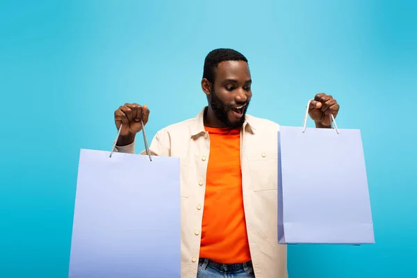 Здивований афроамериканський чоловік тримає сумки ізольовані на синьому — стокове фото