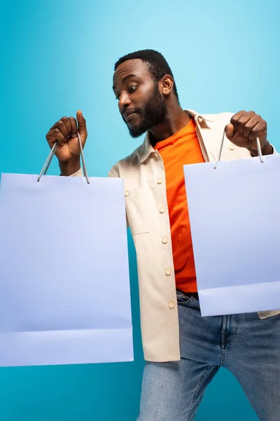 Curioso homem americano africano olhando em saco de compras isolado em azul — Fotografia de Stock