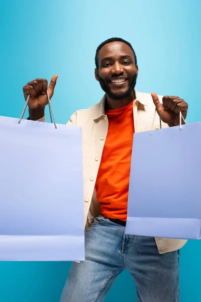 Alegre afroamericano hombre sosteniendo bolsas de compras mientras mira a la cámara en fondo azul - foto de stock