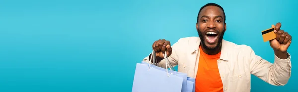 Stupito uomo afroamericano che mostra carte di credito e borse della spesa isolate su blu, banner — Foto stock