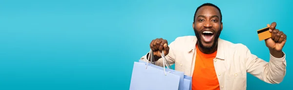 Étonné homme afro-américain montrant carte de crédit et sacs à provisions isolés sur bleu, bannière — Photo de stock