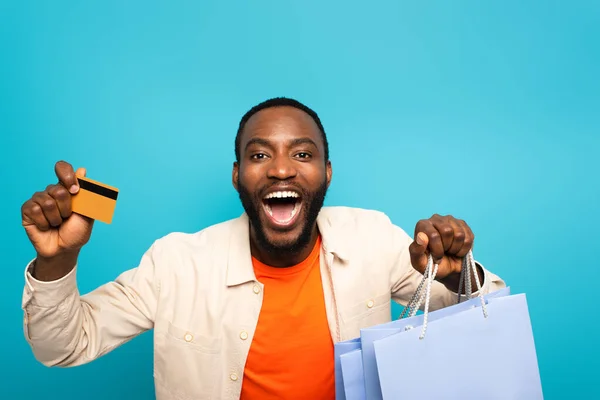 Homme afro-américain étonné avec la bouche ouverte montrant les achats et la carte de crédit isolé sur bleu — Photo de stock