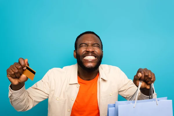 Hombre afroamericano emocionado con tarjeta de crédito y bolsas de compras sonriendo con los ojos cerrados aislados en azul - foto de stock