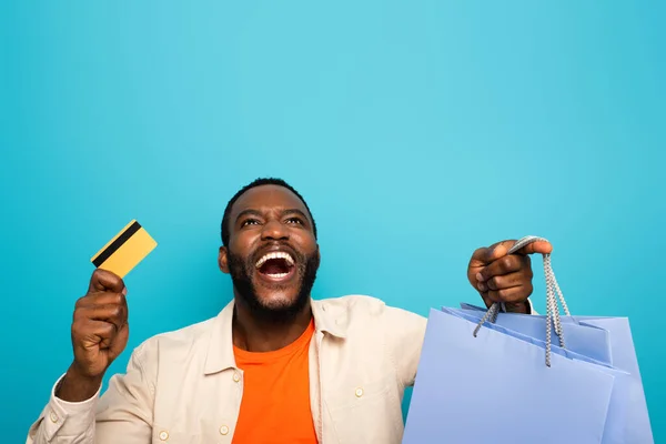 Étonné homme afro-américain avec des sacs à provisions et une carte de crédit en regardant isolé sur bleu — Photo de stock