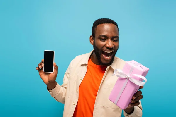 Изумленный африканский американец с подарочной коробкой и смартфоном с пустым экраном, изолированным на голубом — стоковое фото