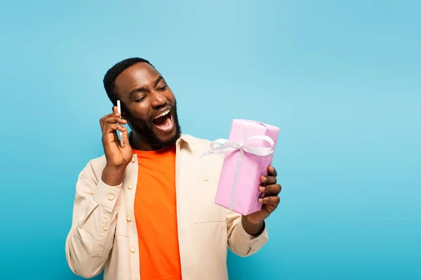 Astonished african american man looking at present while talking on smartphone isolated on blue — Stock Photo