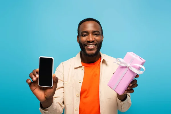 Smiling african american man holding present and smartphone isolated on blue — Stock Photo