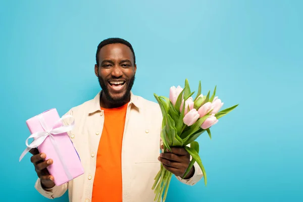 Felice uomo africano americano con scatola regalo e tulipani ridere della fotocamera isolata sul blu — Foto stock
