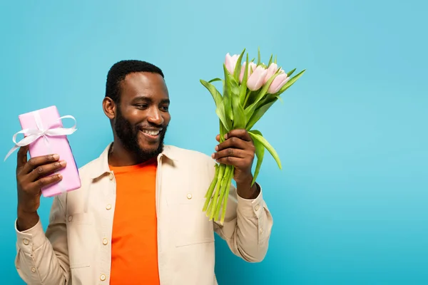 Gioioso afroamericano uomo in possesso di scatola regalo e tulipani isolati su blu — Foto stock