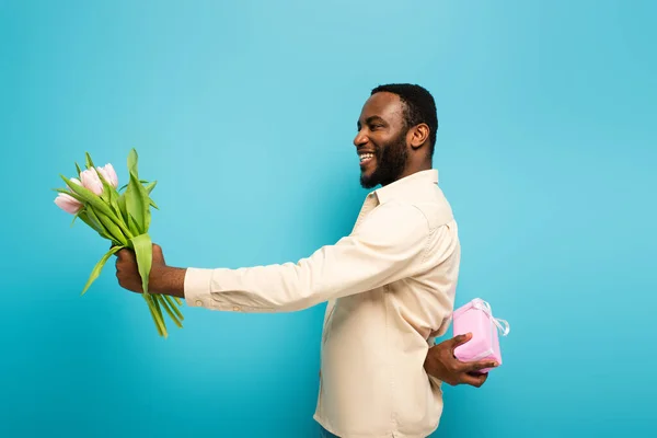 Gioioso afro-americano uomo che presenta tulipani mentre tiene la scatola regalo dietro la schiena su sfondo blu — Foto stock