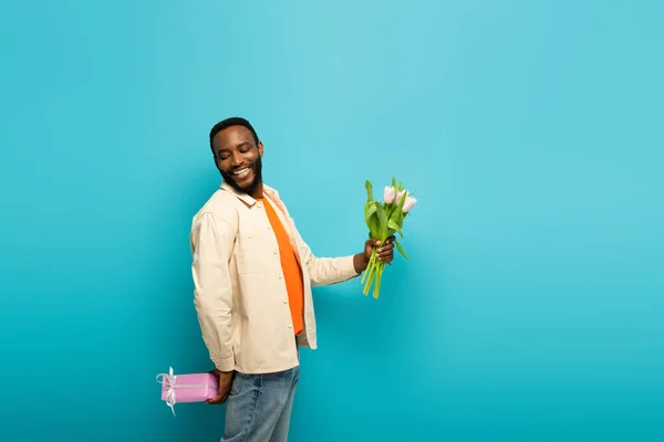Веселий афроамериканський чоловік тримає тюльпани і ховає подарунок позаду на синьому — стокове фото