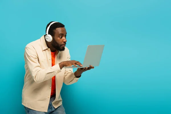 Surpreso homem americano africano em fones de ouvido usando laptop em fundo azul — Fotografia de Stock