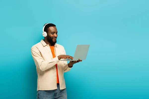 Pleased african american man in headphones typing on laptop on blue background — Stock Photo