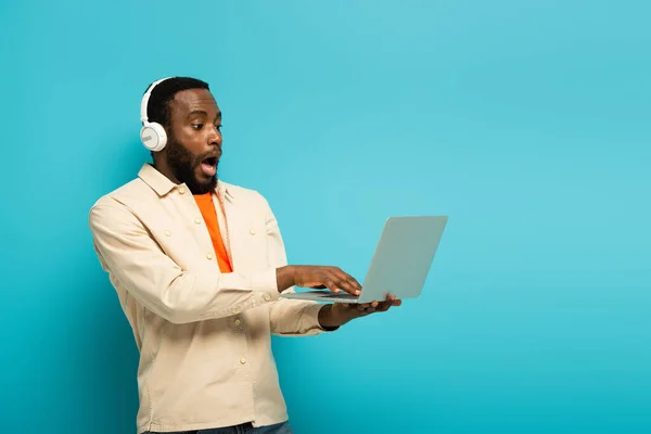 Homme afro-américain choqué dans les écouteurs à l'aide d'un ordinateur portable sur fond bleu — Photo de stock