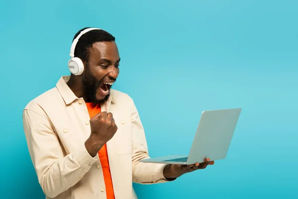 Excité homme afro-américain dans les écouteurs montrant geste de victoire tout en tenant ordinateur portable sur bleu — Photo de stock