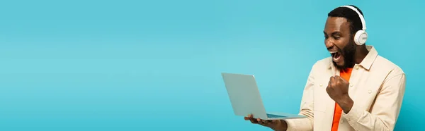 Uomo americano eccitato con cuffie e laptop mostrando gioire gesto isolato su blu, banner — Foto stock
