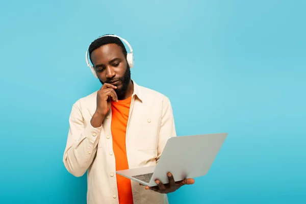 Hombre afroamericano pensativo en auriculares mirando portátil sobre fondo azul - foto de stock