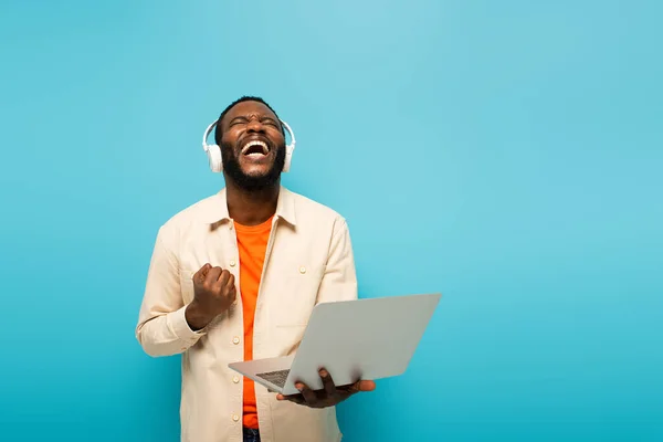 Hombre afroamericano emocionado en auriculares que muestran gesto de éxito mientras está de pie con el ordenador portátil aislado en azul - foto de stock