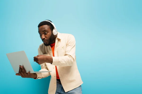 Hombre afroamericano enojado en auriculares que muestran el puño cerrado cerca de la computadora portátil sobre fondo azul - foto de stock